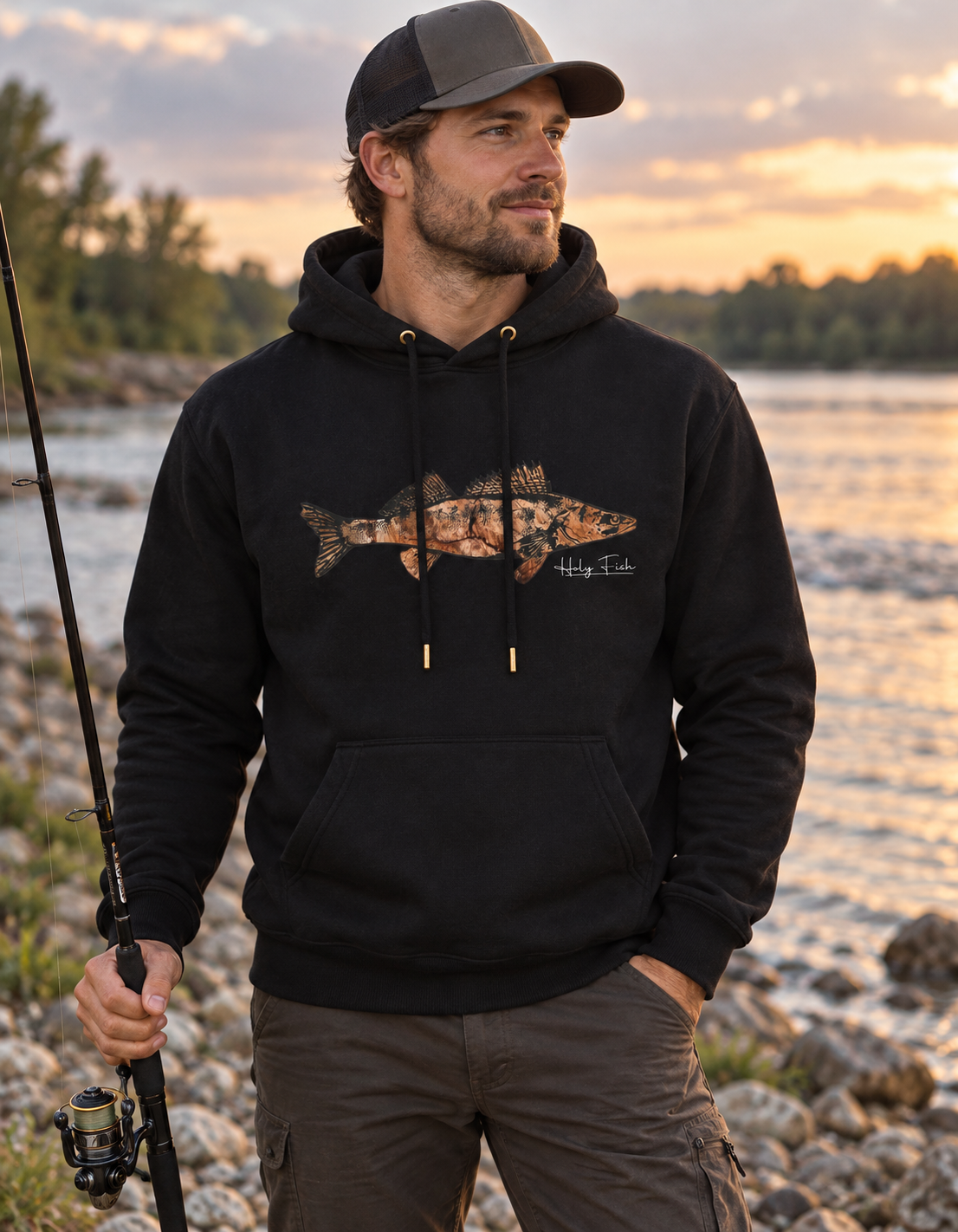 Angler am Fluss trägt schwarzen Holy Fish M13 Zander-Hoodie mit kupferfarbenem Fischmotiv