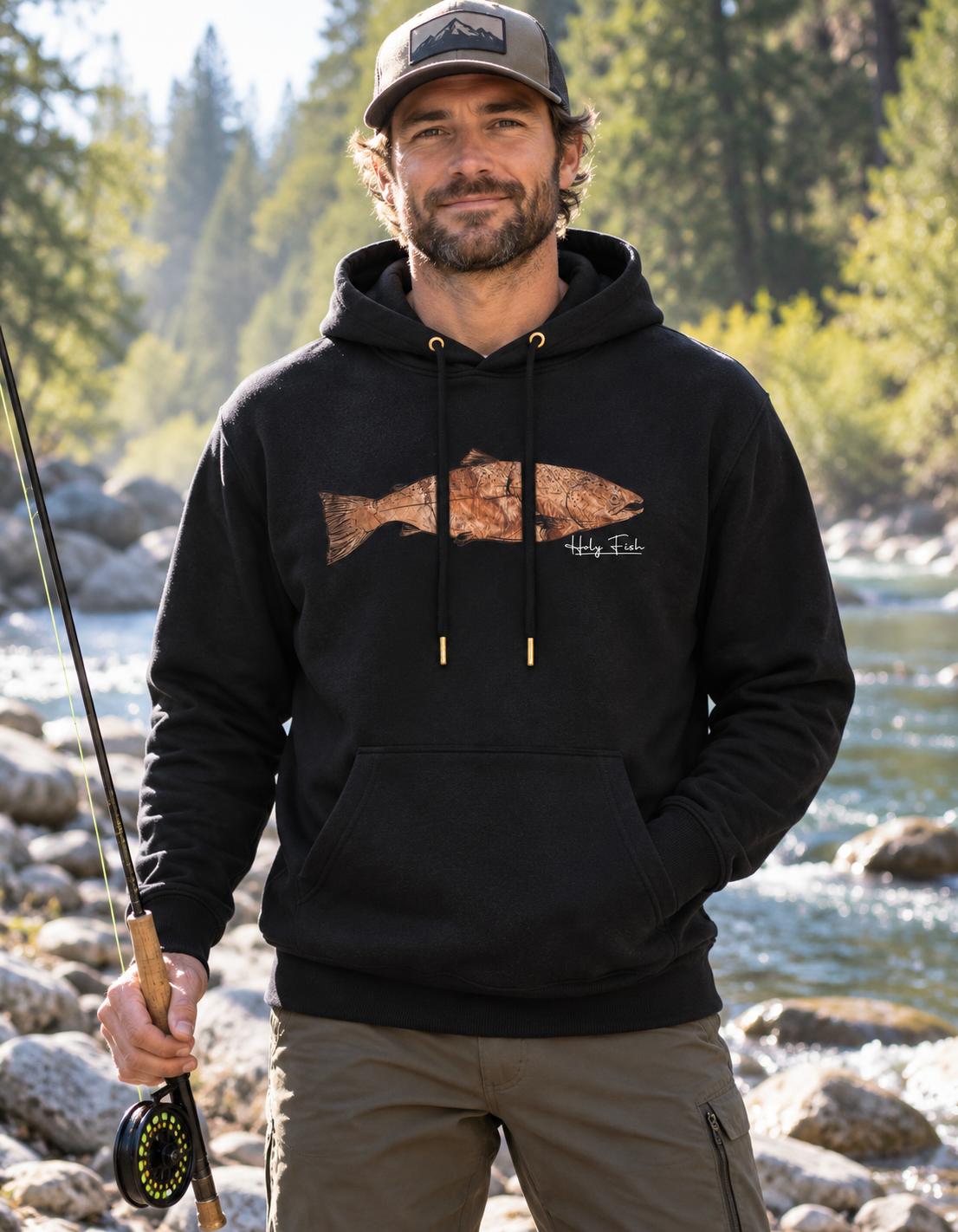 Angler am Fluss trägt schwarzen Holy Fish M11 Forellen-Hoodie mit kupferfarbenem Fischmotiv