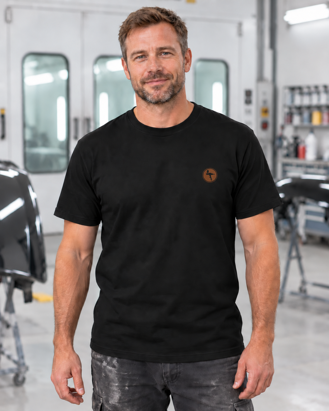 Erwachsener Mann trägt das schwarze Workaholicz Lackierer T-Shirt mit kleinem Lackierpistolen-Brustmotiv in einer Lackierwerkstatt.