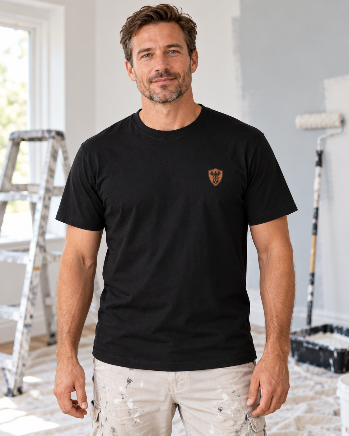 Erwachsener Maler trägt das schwarze Workaholicz T-Shirt mit kleinem Adler-Brustwappen in einem Raum mit Malerwerkzeug