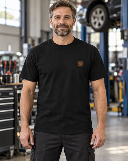 Erwachsener Mann trägt das schwarze Workaholicz Mechatroniker T-Shirt mit kleinem Zahnrad-Brustwappen in einer Werkstatt